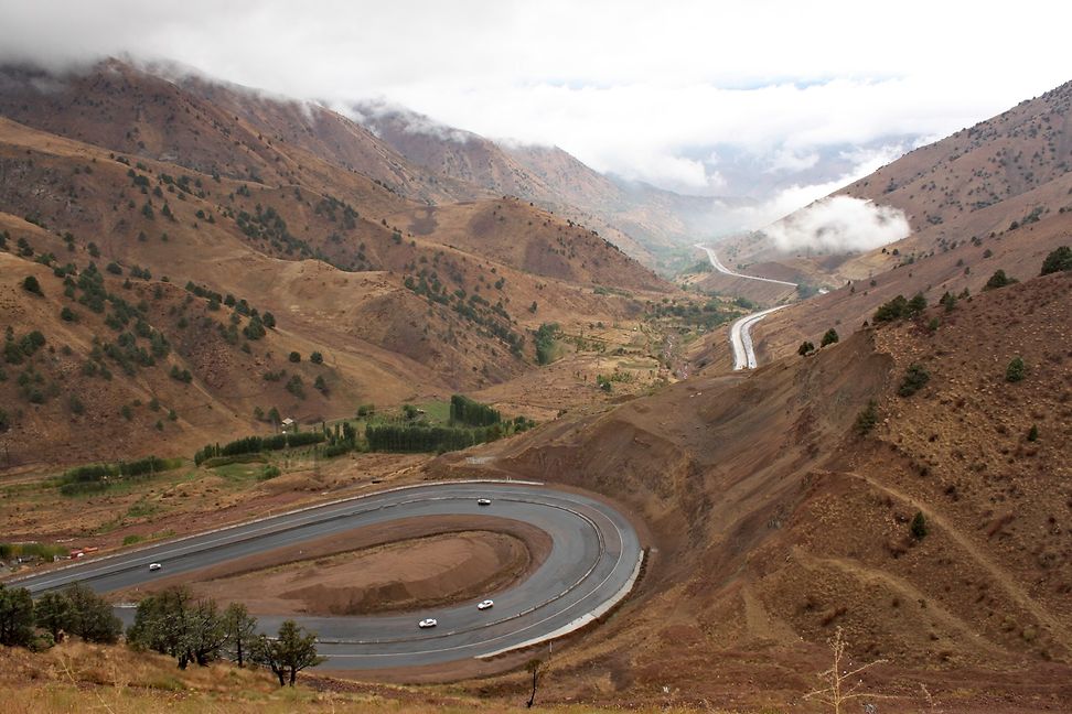 In a narrow valley between brown and cloud-covered mountain slopes runs a winding road on which a few cars are travelling.