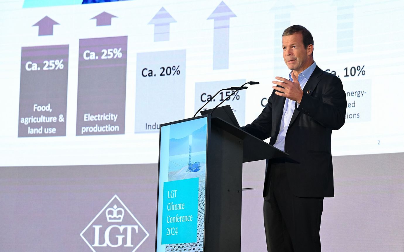 A middle-aged man with dark hair stands at a lectern and speaks. 