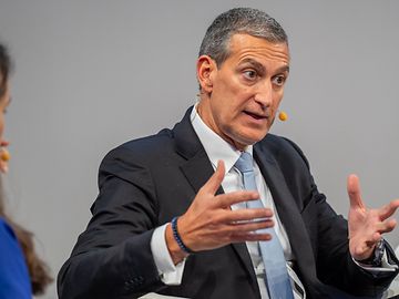 A middle-aged man with dark grey hair, wearing a dark suit, white shirt and light blue tie