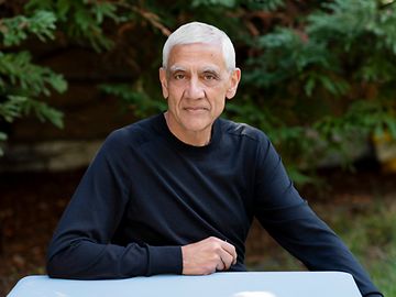 A casually dressed man with white hair is sitting outside in front of fir trees, his arm resting on the table in front of him.