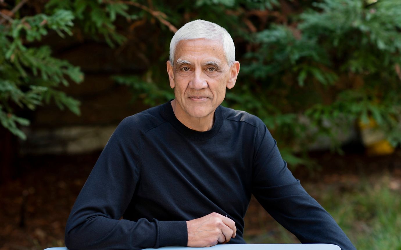 A casually dressed man with white hair is sitting outside in front of fir trees, his arm resting on the table in front of him.