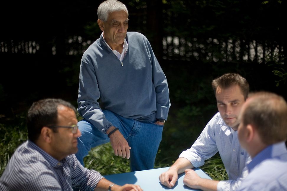 Four casually dressed men are chatting at an outdoor table - one man is standing.