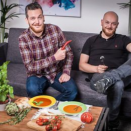 Zwei junge Männer sitzen auf einer grauen Couch, vor ihnen auf dem Tisch stehen zwei Suppenteller und etwas Gemüse. 