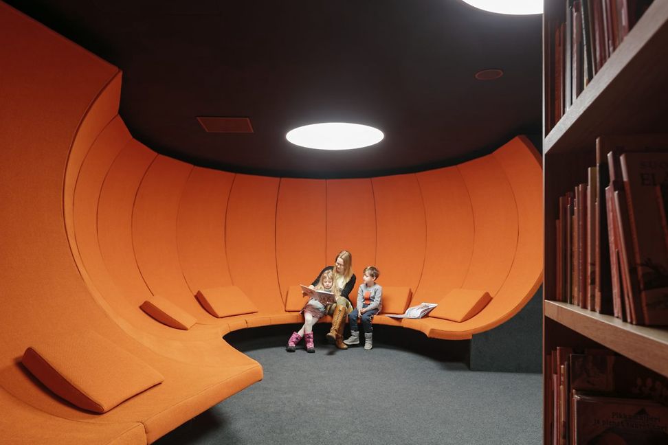 In an enclosed room with high orange seats, a woman reads a book to two children.