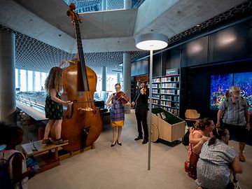 Three string musicians play in a high, open library. A bookshelf, an aquarium, and people pose for a photo in the background.