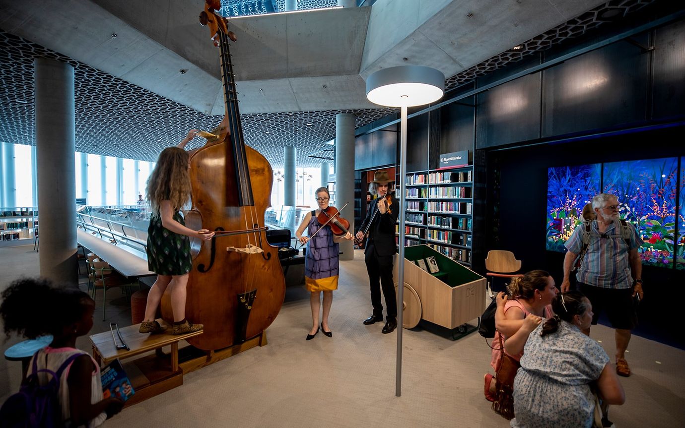 Three string musicians play in a high, open library. A bookshelf, an aquarium, and people pose for a photo in the background.