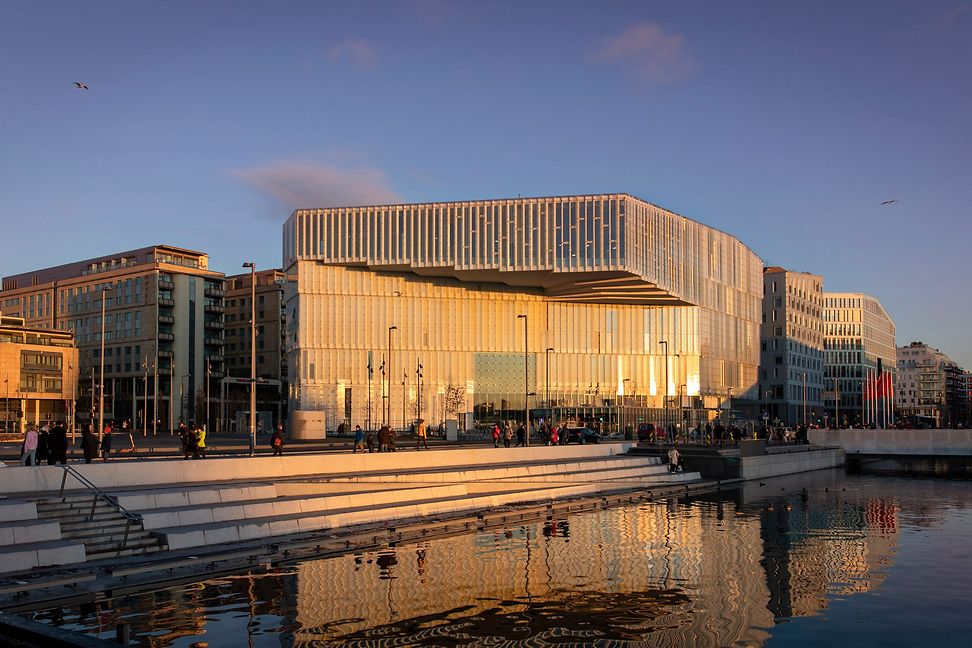 A large building with a glass façade and vertical louvres. A wide staircase leads from the forecourt with people to the water.