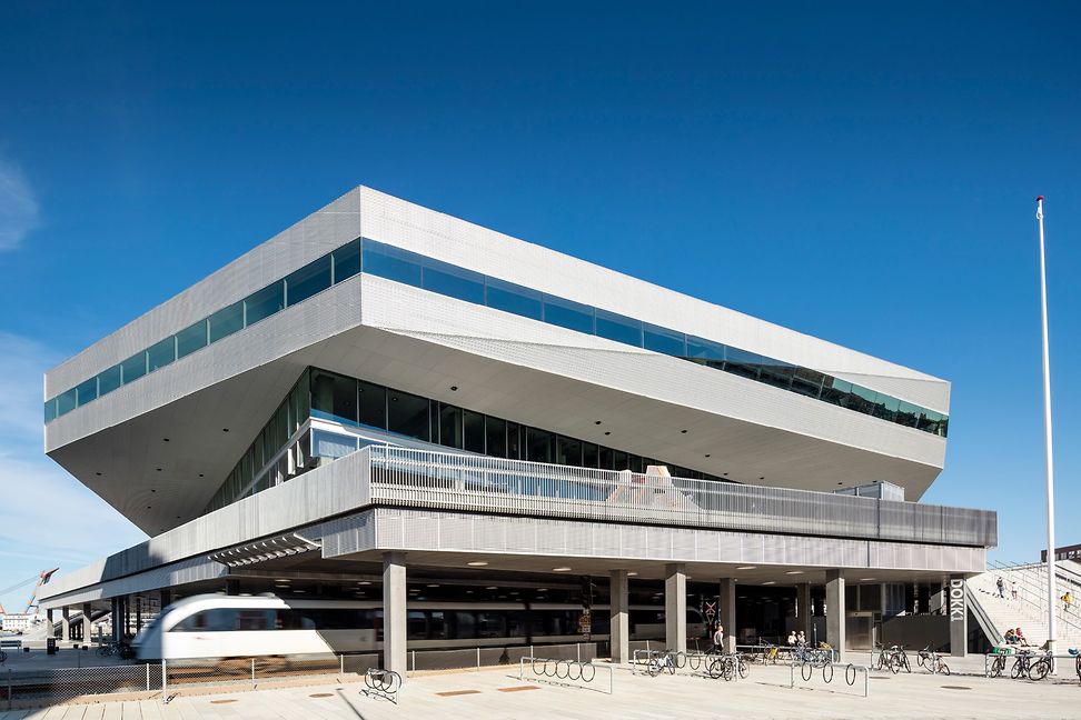 A large modern building with staggered levels and a large forecourt with bike racks and bicycles. A train passes underneath.