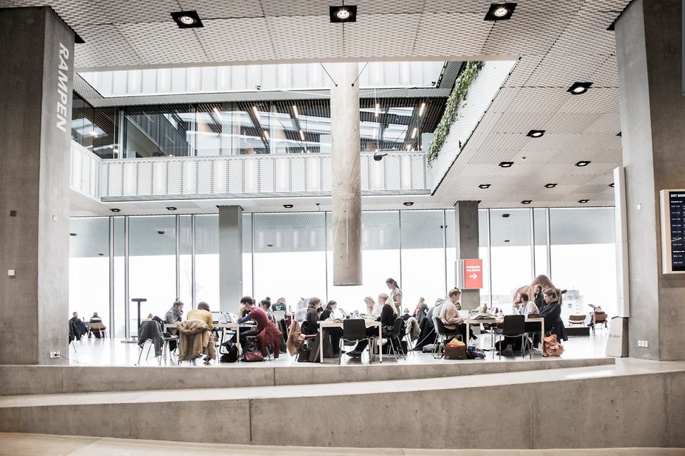 Many people are sitting at tables in a bright, high-ceilinged room. A large metal tube hangs from the ceiling above them.