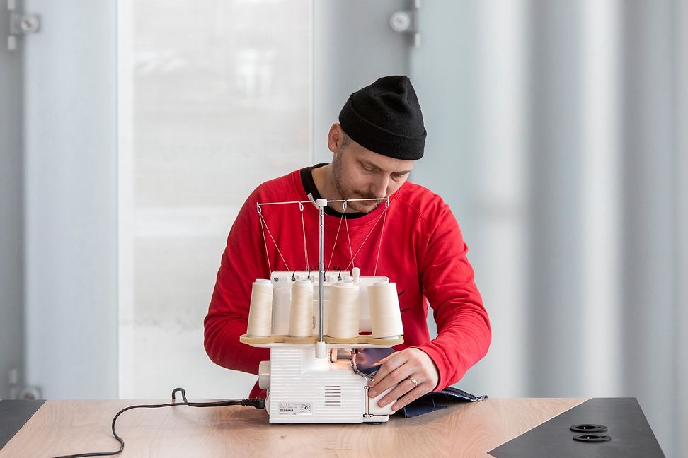 Ein junger Mann mit schwarzer Mütze und rotem Oberteil bedient eine Nähmaschine mit beigen Garnrollen.