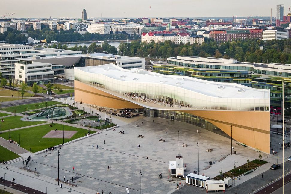 Modernes, langgezogenes Gebäude mit Vorplatz und Park. Die untere Hälfte aus Holz, die obere aus Glas mit einer Terrasse.