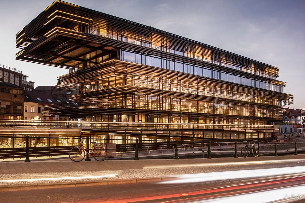 A large modern building lit at dusk with a glass facade interspersed with horizontal louvres in an urban street setting.