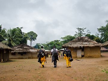 Three men with rucksacks, two in yellow and black work clothes, walk through a village of thatched huts