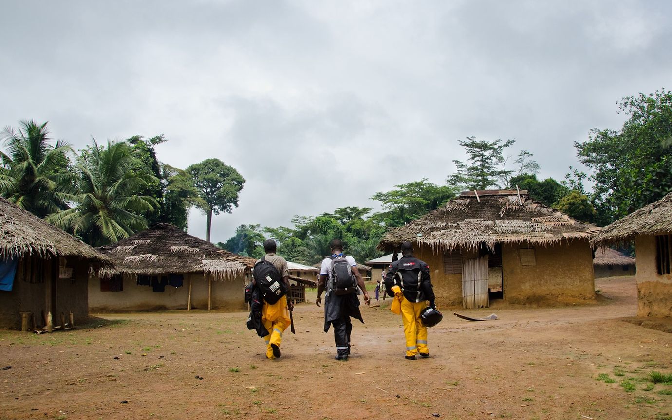 Three men with rucksacks, two in yellow and black work clothes, walk through a village of thatched huts