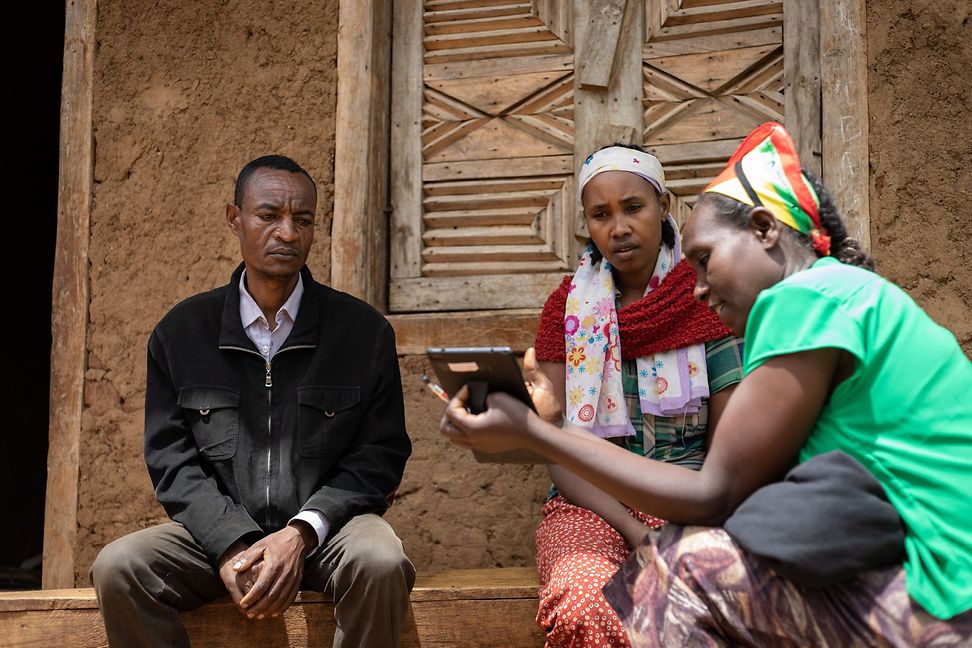 A man and two women are sitting in front of a mud house. One woman holds a tray and shows the others something on it.