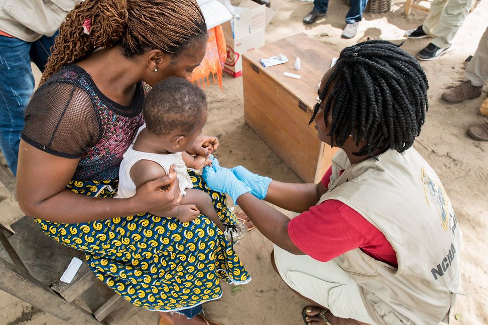 A child sits on a woman's lap. Another woman, wearing blue gloves, is squatting in front of them and treating the child's hand.