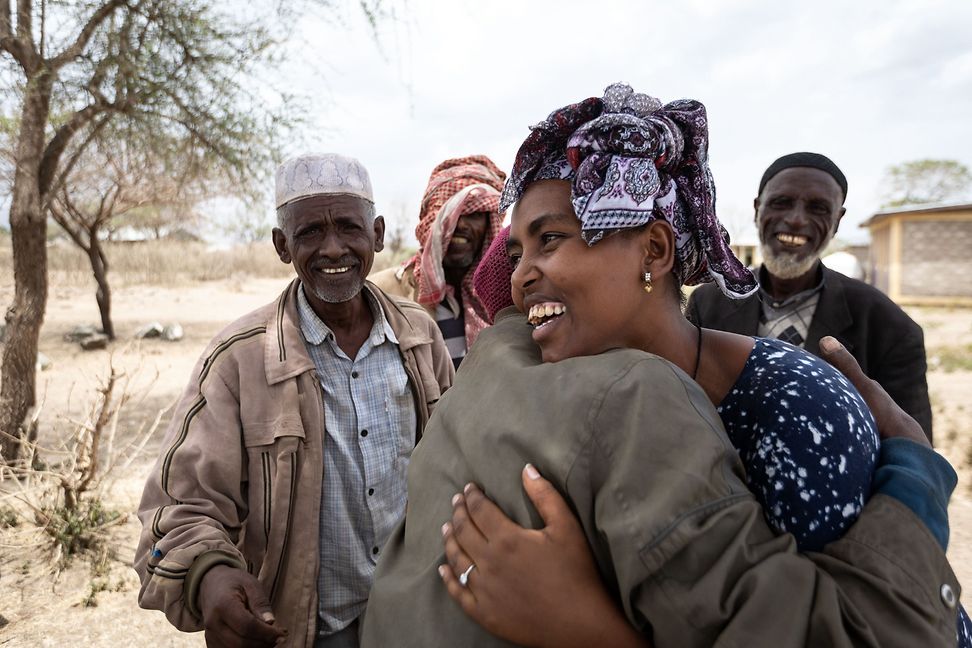 A woman hugs another person, both laughing. Three men laugh behind them. They stand in a barren setting with bare trees.