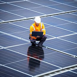 Ein Mann in gelber Jacke, blauer Hose und mit weissem Helm hält einen Laptop und kauert dabei inmitten von Solarmodulen.