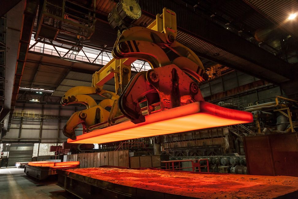 A massive mechanical device lifts red-hot molten metal over a workbench in an industrial environment.