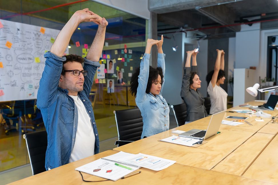 A group of people is sitting at a meeting desk, stretching their arms overhead with eyes closed. 