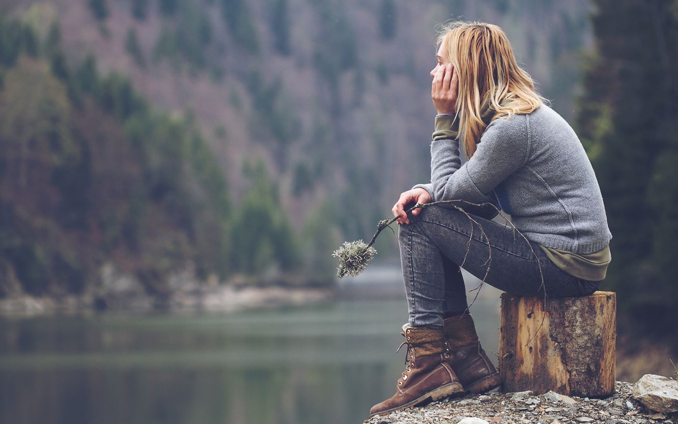 In einer herbstlich, kargen Berglandschaft sitzt eine junge Frau in bedrückter Körperhaltung auf einem Holzblock.