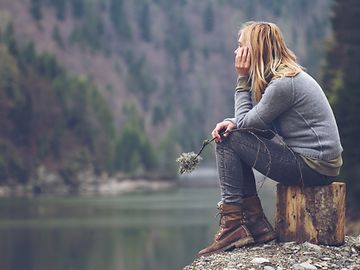 In an autumnal, barren mountain landscape, a young woman is sitting on a block of wood in a depressed posture.