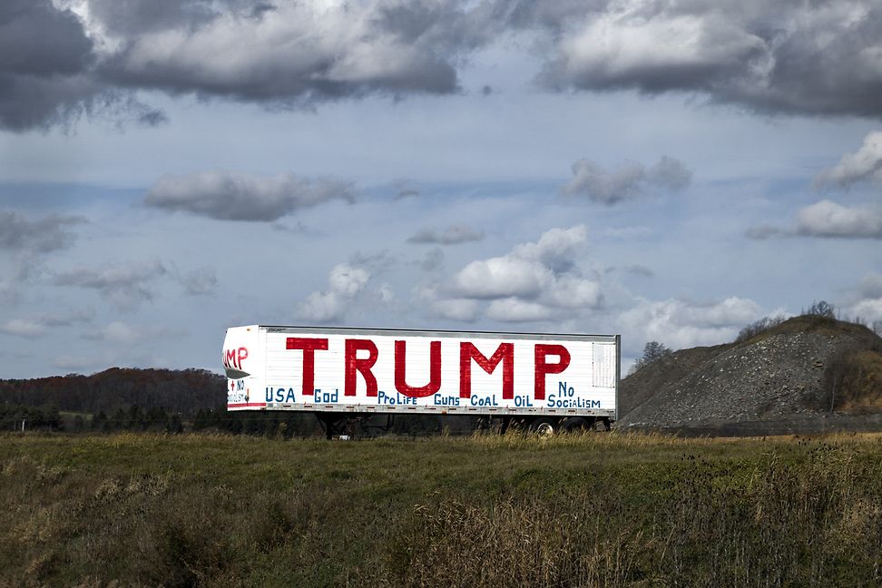 In a rural field, a giant billboard advertises Trump in large letters, with a blue sky in the background.