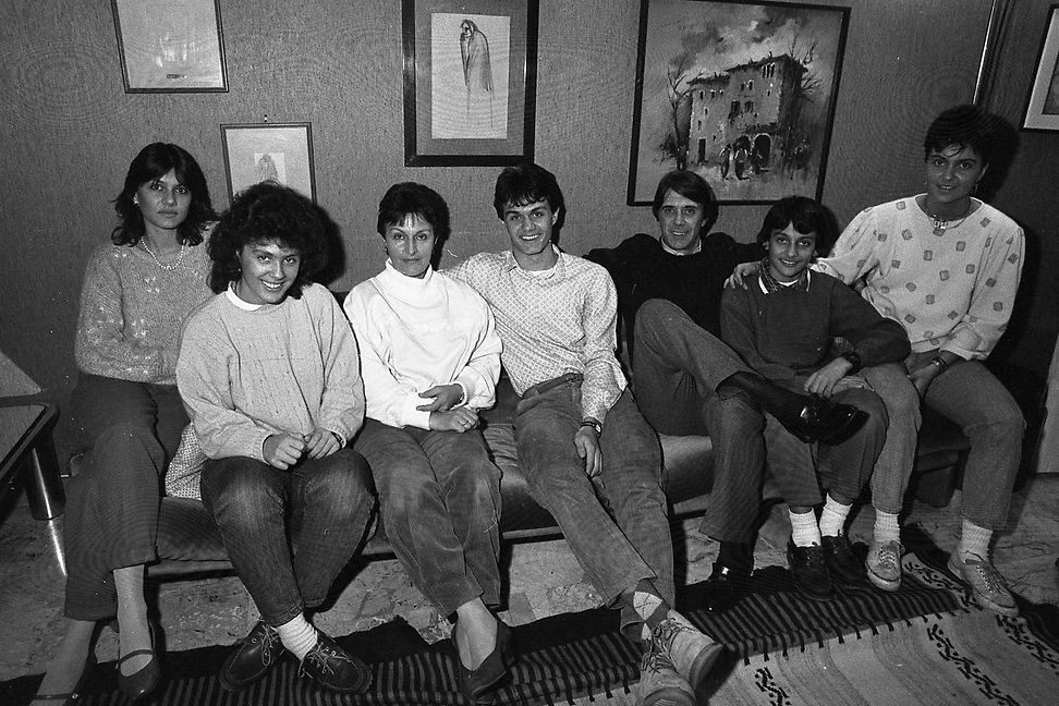 Seven people, two middle-aged and five younger, are sitting on a sofa in front of a wall with pictures.