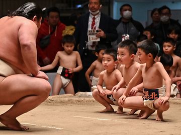 Ein Sumoringer kniet vor drei kleinen Jungen, ebenfalls in traditioneller Sumobekleidung. Im Hintergrund stehen Zuschauer.