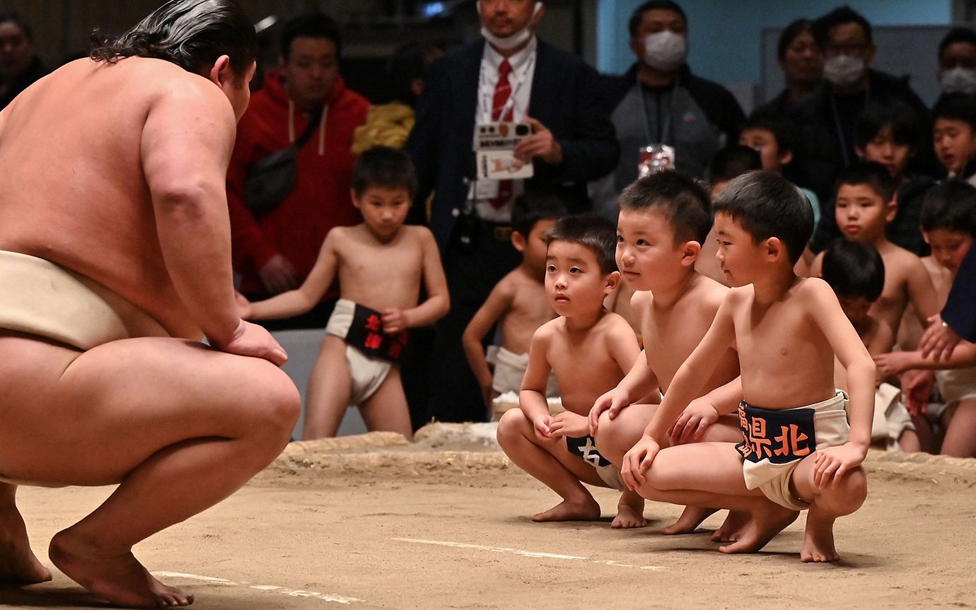 A sumo wrestler kneels in front of three young boys, also dressed in traditional sumo clothing. Spectators in the background.