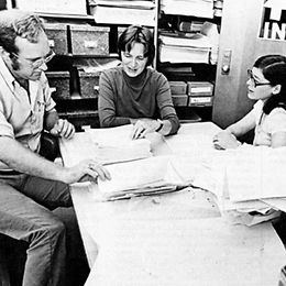 Historic black-and-white photograph of two women and a man in an office setting, reviewing documents and engaging in discussion.