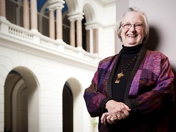 Eine alte Frau in dunkelrotem und violettem Kleid posiert in einer Säulenhalle und lächelt in die Kamera.