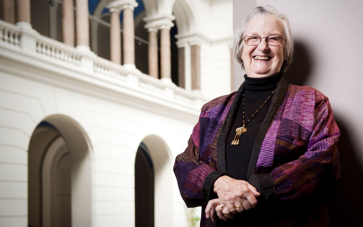 An old woman in a dark red and purple dress poses in a portico, smiling at the camera.