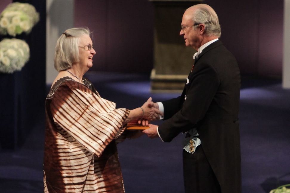 A woman in a patterned dress receives an award from a man at a royal court, surrounded by an audience.