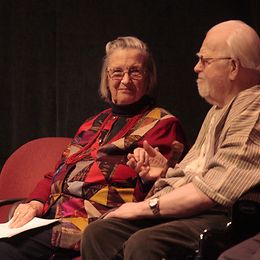 An elderly woman and man sit side by side, exchanging a thoughtful glance as the woman holds a document in her hand.