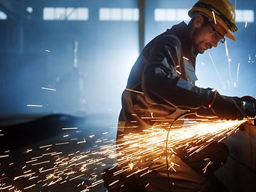 A man in protective gear works at a workbench in a factory setting, with welding sparks flying as he works.