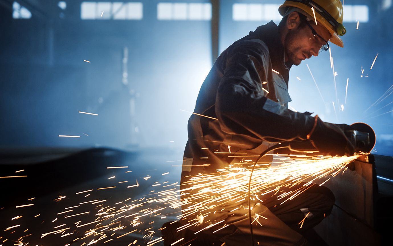 A man in protective gear works at a workbench in a factory setting, with welding sparks flying as he works.