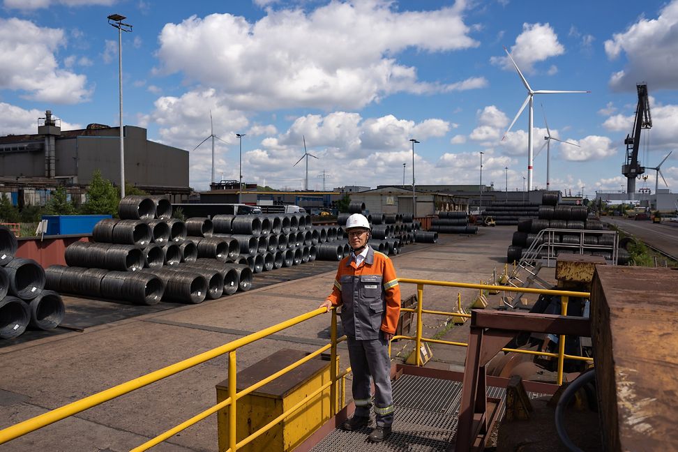 A man in workwear stands outside on a factory site, with wind turbines spinning in the background.