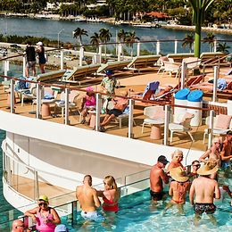 Viele Menschen befinden sich bei sonnigem Wetter auf dem Deck eines grossen Schiffes, einige im Pool, andere in Liegestühlen.