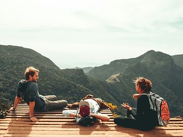 Drei jüngere Personen in Freizeitkleidung sitzen auf einem Holzsteg mit Blick auf bewaldete Berge.