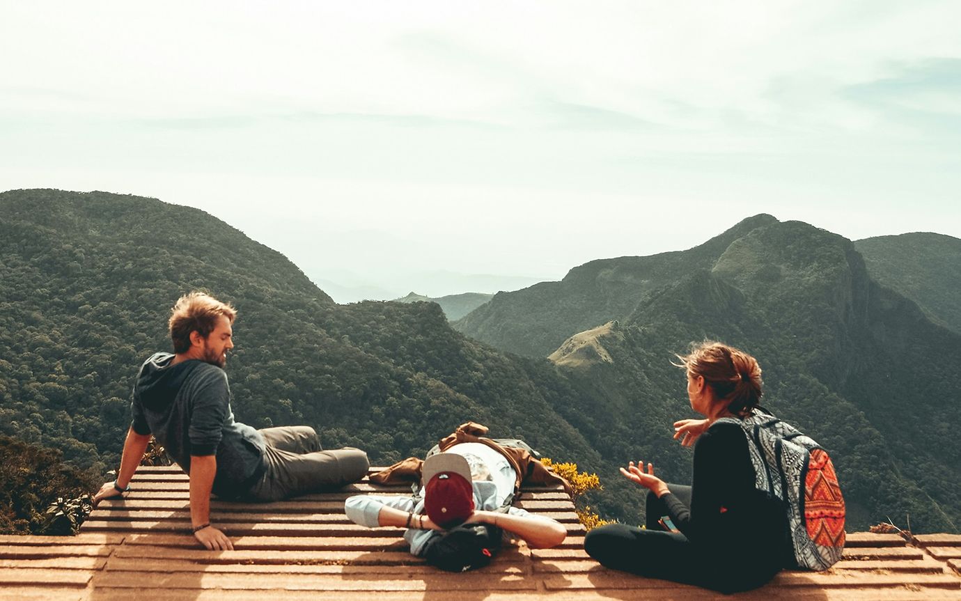 Three young people in casual clothes sit on a wooden footbridge with a view of wooded mountains.