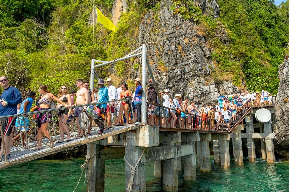 Many people in leisure and swimwear walk along a wooden walkway over clear water and in front of a rocky, wooded cliff.