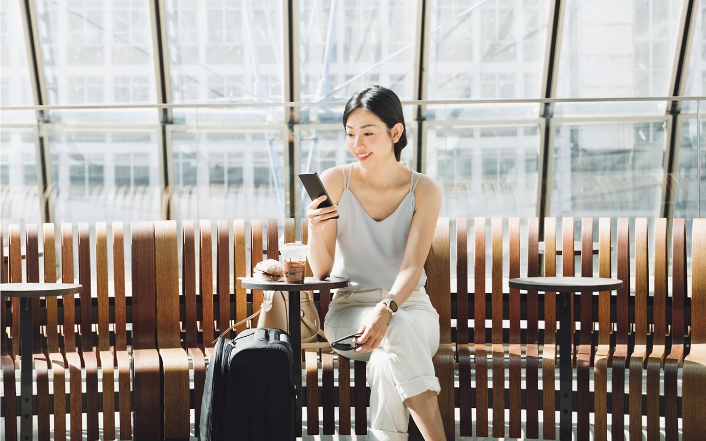 A woman sits on a bench in a bright, sunlit room with large windows, smiling as she reads.