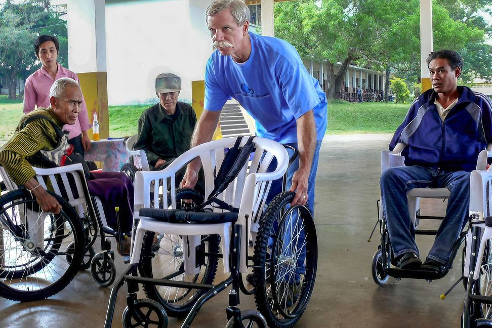 A diverse group of people are sitting in wheelchairs made from a plastic chair, in the center a nurse is pushing 