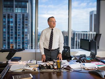 A man in a tie stands behind a desk in an office, documents laid out in front of him