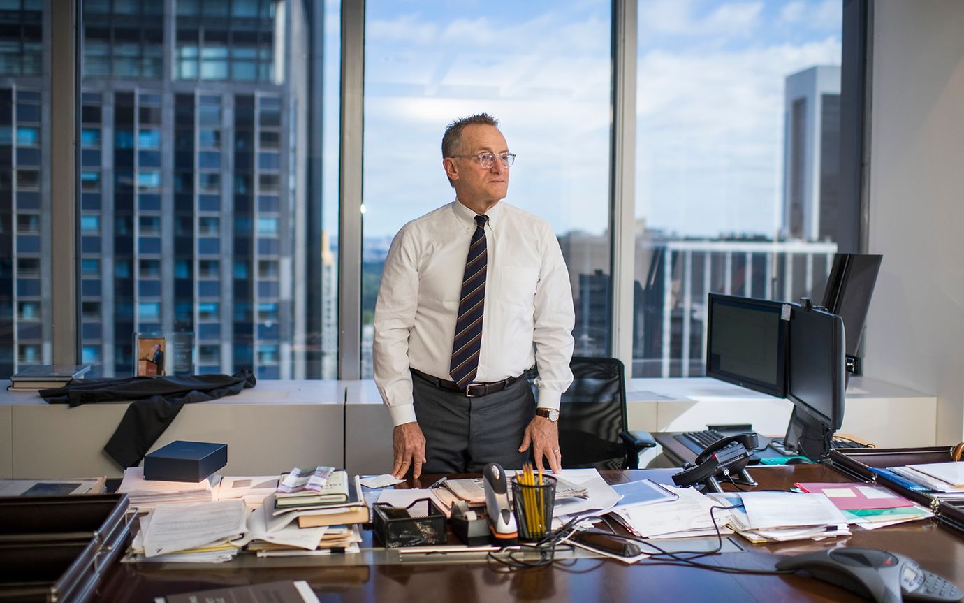 A man in a tie stands behind a desk in an office, documents laid out in front of him