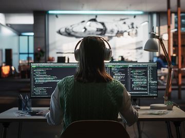 A person, seen from behind, sits wearing headphones in front of multiple screens in a dimly lit office.