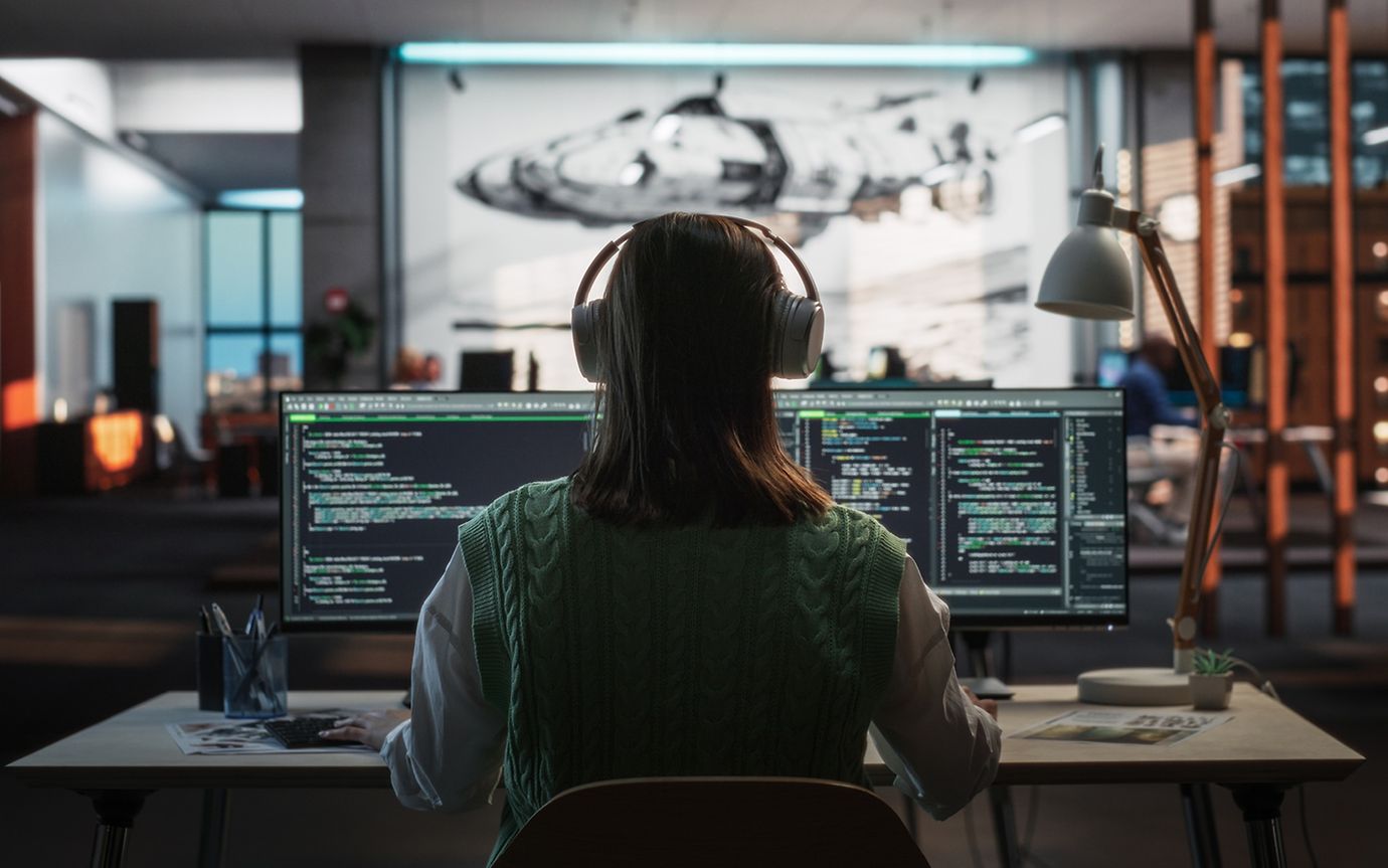 A person, seen from behind, sits wearing headphones in front of multiple screens in a dimly lit office.