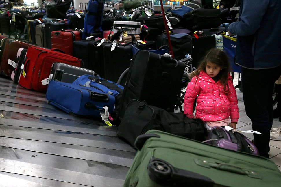 A child watches as large suitcases pile up on a conveyor belt, while many people with large luggage trolleys wait nearby.