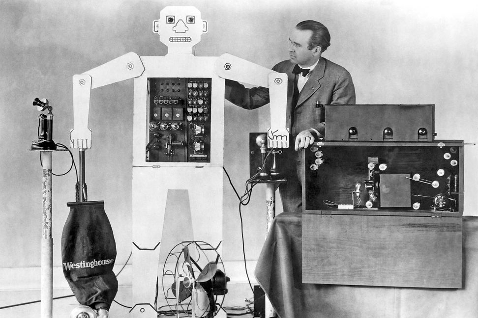 A human-sized figure with a face and moving limbs stands next to a man and a box with cables and controls.